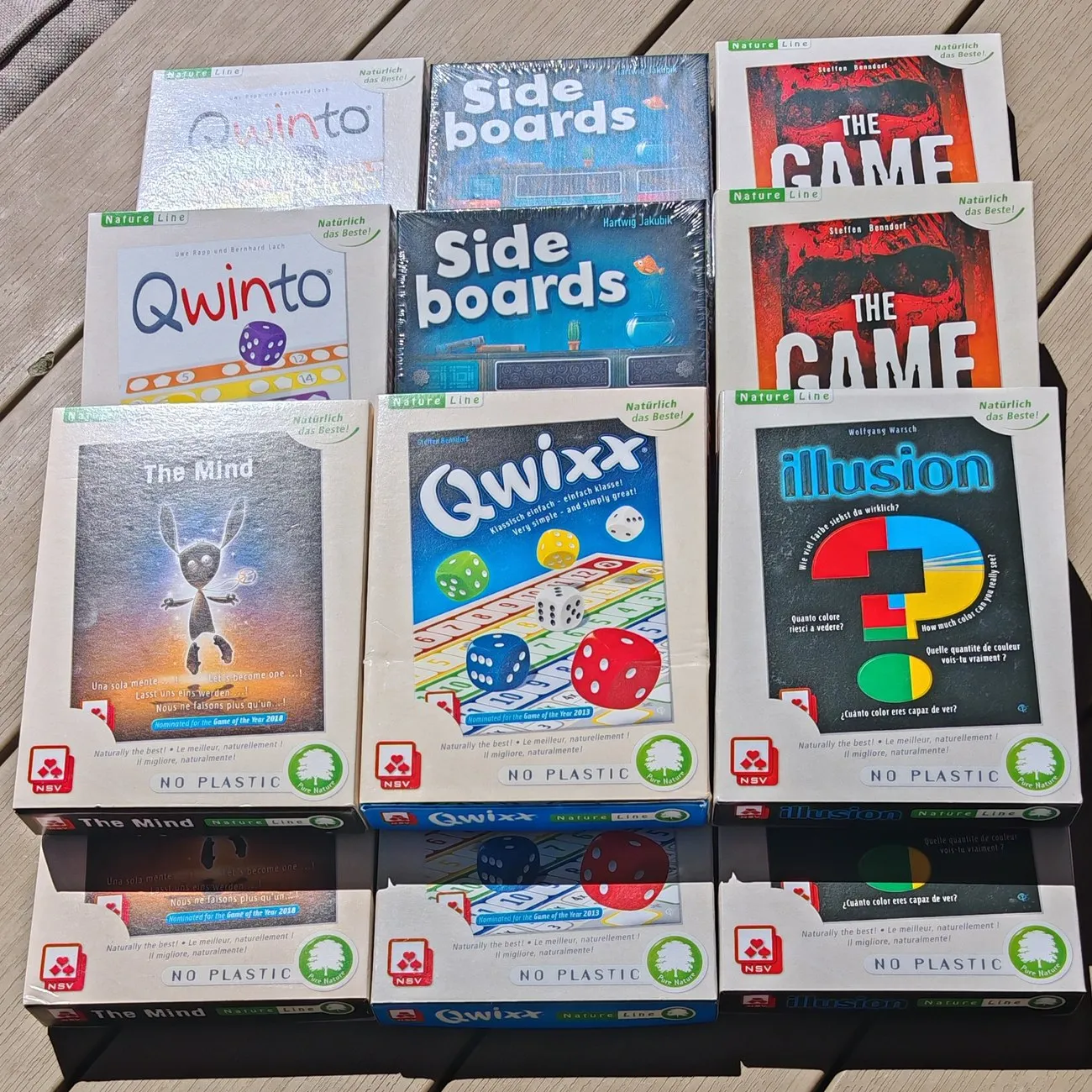 Zwölf Spieleschachteln des Nürnberger-Spielkarten-Verlags aus der Natureline-Reihe, ausgelegt auf einer Holzterrasse